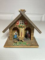 Front view of the vintage wooden weather house showing the roof, central thermometer, and figurines in the side alcoves with deer figures at the front.