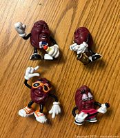 Four California Raisins raisin figurine toys on wood surface showing front details, poses, and accessories like microphone, guitar, and saxophone.