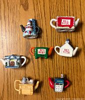 Top down photo showing all 7 mini ceramic tea pots with various Red Rose designs, each uniquely styled including buildings, stoves, and calendar elements.