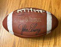 Close-up photo of a Wilson football showing the John Elway signature and Wilson logo on the brown textured surface with white stripes and laces.
