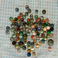 Photo of approximately 70 assorted glass marbles laid out on a blue checkered cloth, showing a variety of colors and patterns.