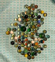 Top-down photo showing approximately 80 assorted glass marbles on a textured blue surface, displaying a variety of colors and patterns including swirls and solid colors.
