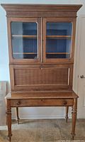 Front view of antique wooden secretary desk with glass doors and shelves in upper cabinet, fold-down writing panel, drawer and turned legs.