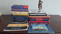 Photo showing an assortment of 18 military-themed books stacked in two piles on a table with a small figurine of a bagpiper on top of one stack. Books cover Canadian military regiments and histories.