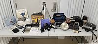 Wide overview of lot on table showing multiple digital and film cameras, projector, lighting, tripod, lenses, boxes and accessories