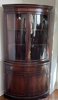 Front view of vintage wooden corner cabinet showing bow front glass doors and lower storage cabinet.