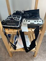 Three braille devices on wooden stand showing Innovision Braille Me, Brailliant BI14, and Braille Pen 12 with cords and carrying cases, plus visible braille cards on lower shelf.