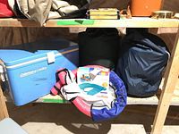 Wide view of wooden shelf with Coleman Lowboy cooler, two sleeping bags, and pool floats placed on shelf and floor beneath.
