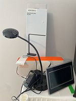 Photo showing the Volvolinc DL2201 smart desk lamp with flexible gooseneck arm, original box, power adapter, and partially visible Logitech Litra Glow box with the Centrios digital photo frame screen visible.