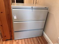 Full front view of gray side load file cabinet with three drawers, lock on top drawer, keys inserted, and hanging files and binders inside.