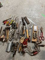 Overview of hand tools spread on floor including screwdrivers, wrenches, saws, hand drills, pliers, scissors, and sharpening stone.