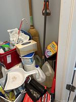 Photo showing various cleaning supplies including white and red buckets, plastic dustpan, broom, mop, and partially used cleaning liquids in plastic bottles and jug.