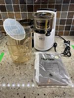 Side view of Hurom juicer main unit, transparent juice collector, and instruction manual in plastic
