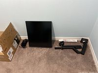 Monitor panel leaning against wall alongside original cardboard box, separate power adapter and disassembled clamp-style arm