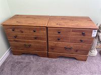Pair of pine veneer nightstands side by side on carpeted floor against light wall, showing front view with all drawers closed