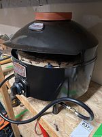 Black metal PizzaQue portable outdoor pizza oven on a wooden surface with hose attached. Visible round terracotta chimney cap on top.