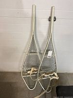 Photo showing the pair of vintage snowshoes featuring white metal frames and metal mesh webbing with canvas foot straps.