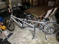 Full view of the Fiori AV Ventura tandem bicycle in silver with two seats and a bike pump placed on the ground nearby.