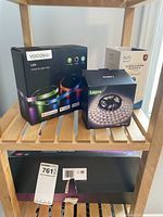 Box packaging of Vocolinc LS3 ColorFlux Light Strip, Eufy indoor security camera, Lepro LED strip, and Philips Shape Light kit on wooden shelf.