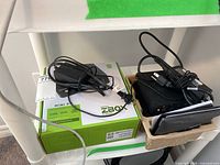 Two Zotac ZBox green retail boxes and brown molded trays holding power adapters, cables, manuals and one mini PC.
