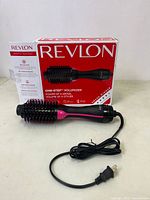 Full lot showing Revlon One-Step Volumizer device in front of original box and product pamphlet on an off-white table surface.
