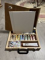 Full view of open wooden paint carrying case showing compartments containing oil paint tubes, small bottle, three canvases propped at back, and wooden palette leaning against case.