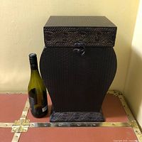 Black metal decorative storage box displayed next to a wine bottle for size reference. Shows curved body and embossed pattern on the top edge and base.