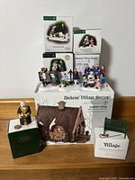 Group shot of six Dickens' Village figurines displayed on boxes including Anglesey Cottage and park bench, showing all items included in lot.