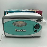 Photo of front facing Hasbro Easy Bake Oven in turquoise and white with box behind it.