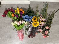 Wide view of the entire collection showing a variety of artificial flowers including tulips, sunflowers, hydrangeas, roses, and other floral types arranged and laid out on the floor.