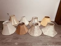 Nine lampshades against a white wall on wood flooring, various shapes and fabric textures visible.
