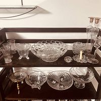 Wide view of vintage crystal and glass items displayed on two black shelves including bowls, dishes, candle holders, and tall candlesticks.