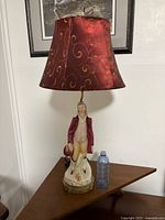 Full view of porcelain figural lamp with red fabric shade and brass base next to a water bottle for scale.