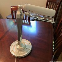 Full view of the adjustable white desk lamp with magnifier panel and base with organizer tray, displayed on wooden table.