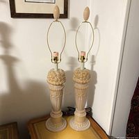 Pair of tall vintage porcelain table lamps with beige glaze and sculpted fruit decoration on top columns, brass fittings and harp, on vintage table surface.