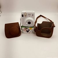 White Fujifilm Instax Mini 11 instant camera, two-part brown faux leather carrying case, and unopened Instax Mini film box stacked beneath the camera.