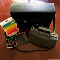 Vintage Polaroid Automatic Land Camera 430 with original black leatherette carrying bag, original user manual, and a Polaroid color stripe card on a wood table.