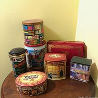 Photo showing all 7 vintage collectible tins arranged on a small wooden table against a cream wall, capturing the variety of shapes and designs including the Werther's Original, Edbury, and Maison Cafe tins.