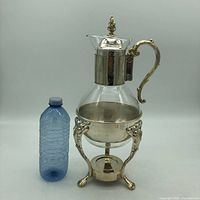 Front view of vintage glass coffee carafe on silver plated ornate three-footed stand. Next to a plastic bottle for size reference.