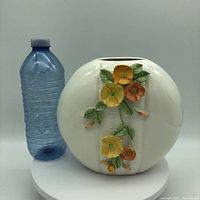 Front view of the white round ceramic vase with colorful bas relief flowers next to a water bottle for scale.