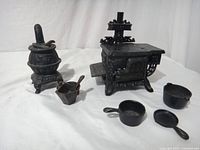 View showing both vintage miniature cast iron stoves, potbelly stove on left with ash bucket/scoop and Queen stove on right with small pots and pans arranged on stove surface.