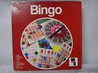 Red square box with Bingo game graphic showing game cards, chips, and spinner on lid.