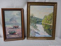 Photo of two vintage original paintings displayed side by side: a riverside landscape in a distressed gold frame and a boats at low tide scene in a brown frame.