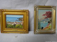 Two framed paintings: left is a painting of Madeira Island on canvas with gold frame, right is a painting of a sailboat on water with pink trees, on hardboard with gold ornate frame.