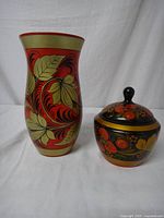 Two wooden Khokhloma items: an 8-inch vase with red, gold, green leaf and floral patterns; and a lidded jar about 4 inches tall with red and gold floral designs on a black background, shown side by side.