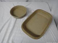 Two beige Pampered Chef stoneware pieces on white fabric background, one round and one rectangular dish.