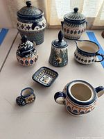 Image showing the assortment of seven pieces of Boleslawiec pottery on a white table including canisters, creamer, sugar bowl, bell, small dish, and miniature shoe figurine.