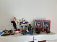 Overview of all items on white shelf including toy bird bank, clown figures, circus clown boxed toy, cast iron elephant, wooden train and carousel penny bank.
