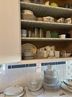 Kitchen shelves with various stacked white plates and bowls with floral patterns, glass tumblers, mugs, and ceramic pitchers.