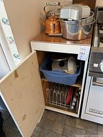 Open kitchen cabinet showing upper shelf with copper kettle, aluminum bowls, and blue plastic bin with pots and mixing bowls.